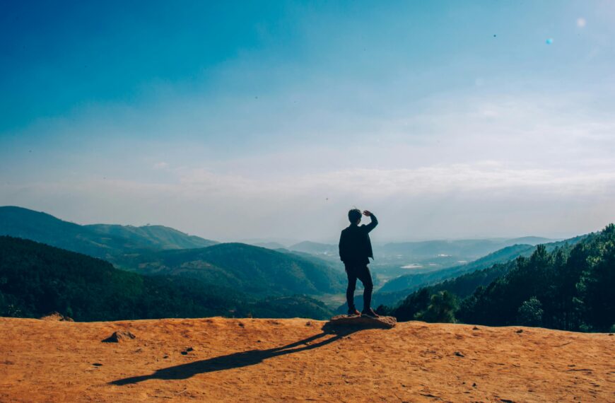 A lone traveler stands on a hilltop, gazing over a vast mountain landscape under a clear blue sky.